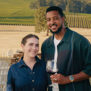 Couple smiling with red wine glasses in front of Stoller Family Estate vineyard rows in Oregon's Dundee Hills